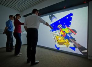 Menschen zeigen auf eine Powerwall im Virtual Reality Labor, auf der ein CAD-Modell zu sehen ist.